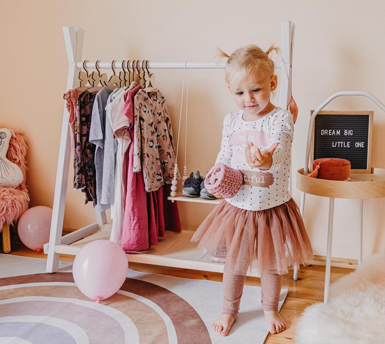 Little girl in a festive dress playing beside a white clothing stand, offering charming inspiration for a toddler room birthday makeover.