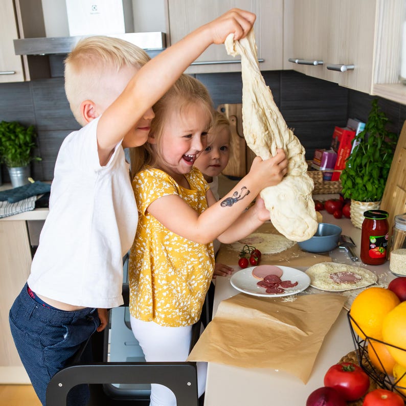 Three children playing with floury pastry dough on a matte black step stool, eliminating unsafe chair climbing at the counter.