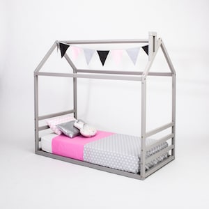 Puede incluir: Una cama de madera gris en forma de casa con un colchón estampado en rosa y gris y una guirnalda de banderas blancas, rosas y negras.