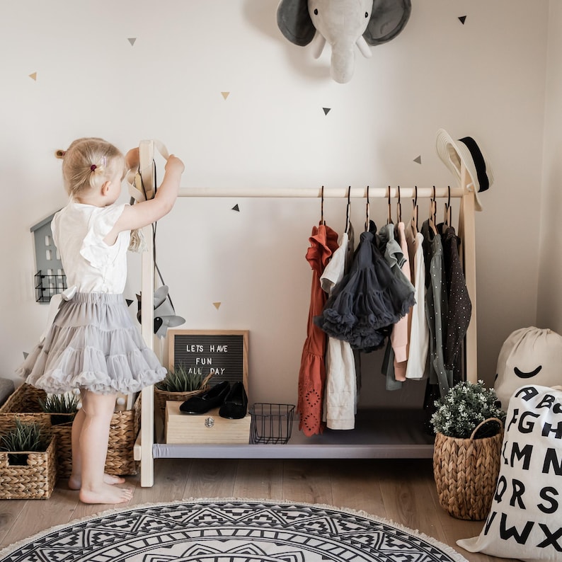 Little girl choosing dresses from a low birch rack, helping foster independent dressing habits in young children before kindergarten.