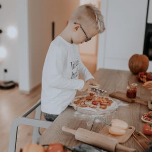 Young boy wearing glasses standing on a slate grey child step stool to make homemade pizza, developing motor skills.