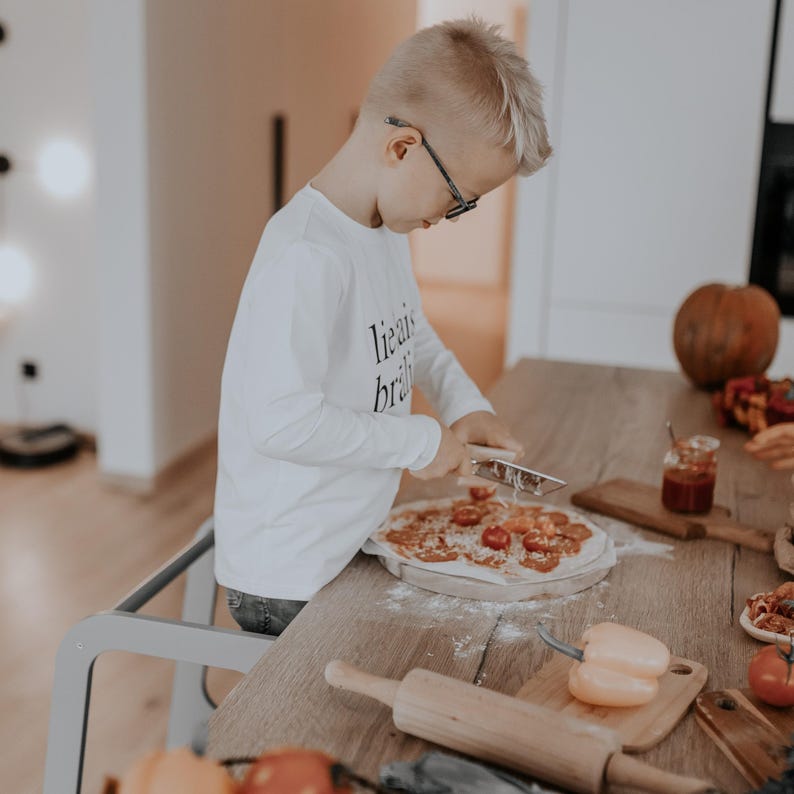 Young boy wearing glasses standing on a slate grey child step stool to make homemade pizza, developing motor skills.