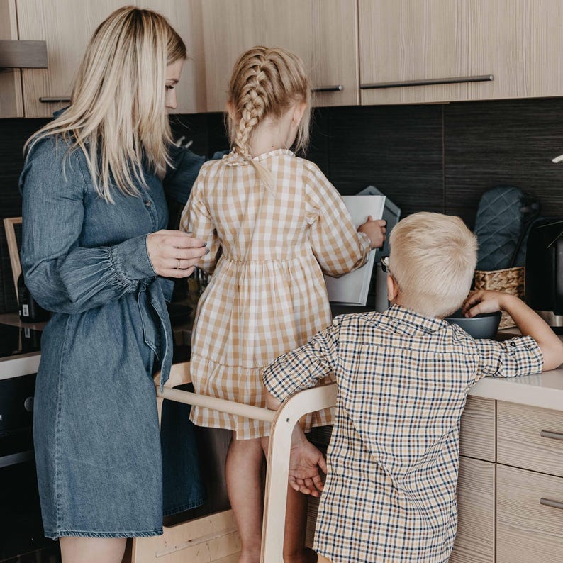 Parents watching their two toddlers stand together on a kitchen counter step stool, making a practical milestone birthday present.