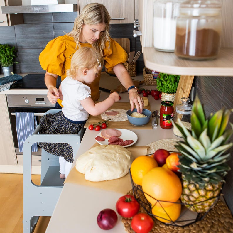 Mother helping her young daughter make fresh snacks on a grey counter booster stool, promoting healthy eating habits.