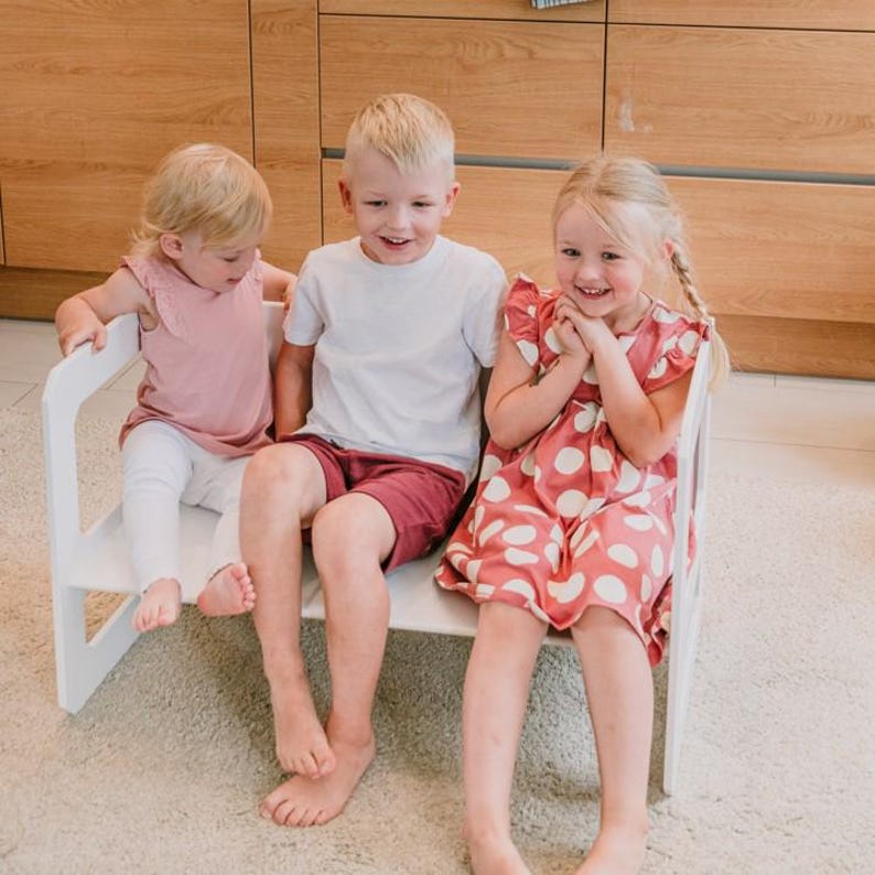Three toddlers sitting side by side on a white wooden activity bench in a nursery.