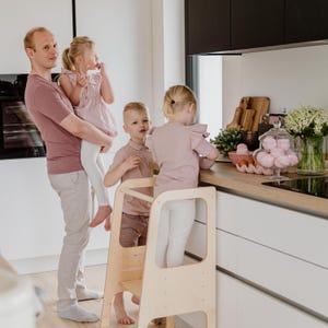 Father holding a toddler next to a standing stool where two older siblings safely help prepare a family meal.