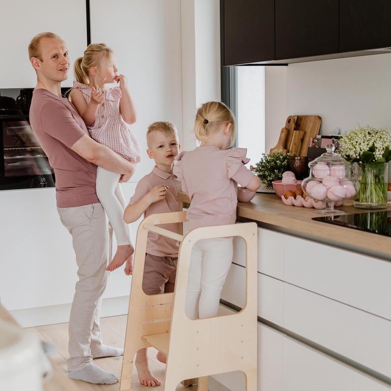 Father holding a toddler next to a standing stool where two older siblings safely help prepare a family meal.