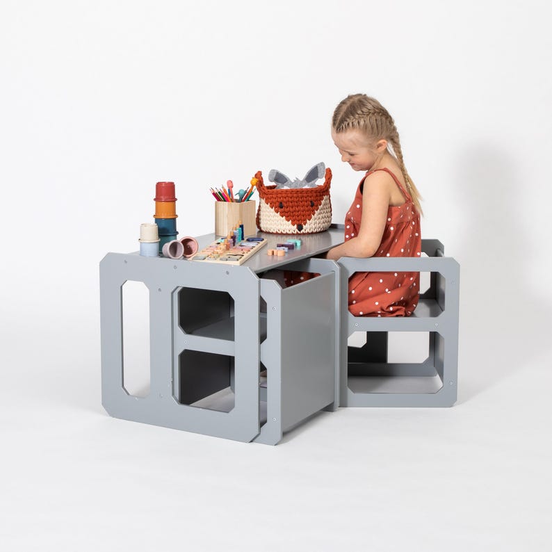 Girl sitting at a grey convertible weaning table with matching cube chairs on a white background.