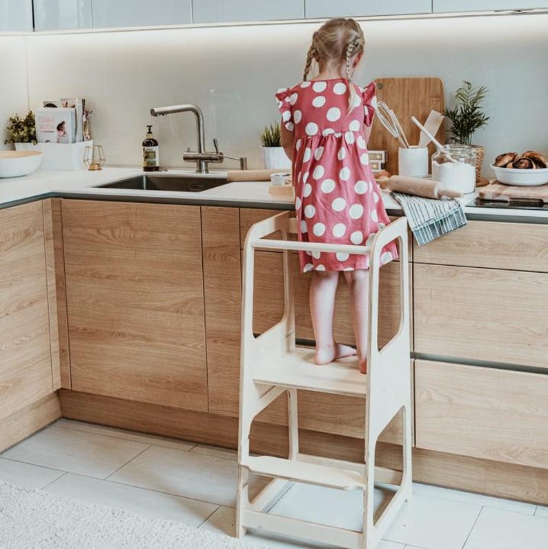 Toddler girl in a polka dot dress standing on a white montessori step stool while reaching for items at the wooden counter.