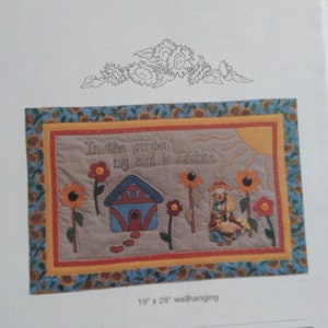 May include: A quilted wall hanging with a beige background featuring a small house, sunflowers, and a person holding a basket. The text "In the garden my soul is sunshine." is stitched in brown thread. The wall hanging measures 19 inches by 29 inches.
