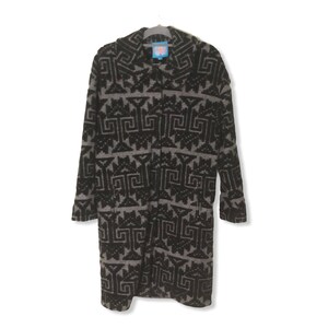 May include: A long, black and gray patterned coat hangs on a hanger. The coat features a geometric design. The coat has a collar and buttons down the front. A small blue tag is visible near the collar.