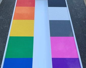 May include: A rectangular white-framed play table with a divided surface. One side features a rainbow of red, orange, yellow, green, blue, and purple Lego-compatible baseplates. The other side has black, white, gray, pink, and purple baseplates.