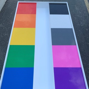 May include: A rectangular white-framed play table with a divided surface. One side features a rainbow of red, orange, yellow, green, blue, and purple Lego-compatible baseplates. The other side has black, white, gray, pink, and purple baseplates.