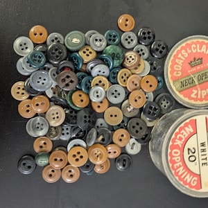 May include: A collection of assorted buttons in various colors, including brown, black, gray, and green. Two vintage spools of Coats & Clark's thread are also present, with labels that read "Neck Opening" and "Zipper". One spool is labeled "20" White".