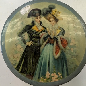 May include: Round, vintage tin with a painted image of a man and woman in 18th-century attire. The man wears a dark coat and hat, while the woman is in a teal dress and feathered hat. The tin has a blue rim and a floral background.