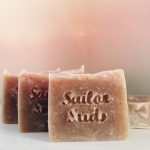 May include: Four bars of handmade soap with a light brown color and a speckled texture. The soap bars have the words "Sailor Suds" stamped on them.