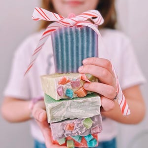 May include: A stack of four colorful handmade soap bars wrapped with red and white striped ribbon. The soap bars have a variety of colors and patterns, including green, purple, and pink.