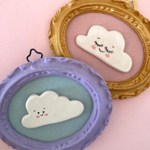Può includere: Due decorazioni a forma di nuvola incorniciate. Una cornice è dorata, l'altra è lavanda. Ogni cornice contiene una nuvola bianca con un volto sorridente e guance rosee. Lo sfondo è rosa chiaro.