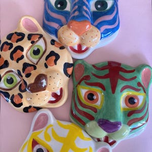 Peut inclure: Quatre décorations murales en céramique représentant des visages d'animaux. Les visages comprennent un tigre bleu avec des rayures roses, un léopard avec des taches noires, un tigre vert avec des rayures rouges et un tigre blanc avec des marques jaunes. Le fond est rose.