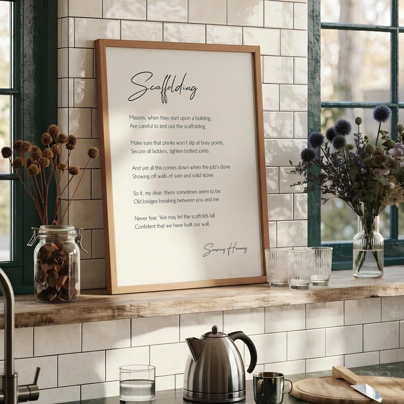 a framed poem titled "Scaffolding" by Seamus Heaney, which is described as a modern and minimalist design. The poem is written in black text on a white background and is framed in a simple wooden frame. The text is arranged in a structured format, with each line beginning with "So, if my dear, there sometimes seems to be" followed by the rest of the poem. The image does not contain any additional text or details beyond the poem itself.