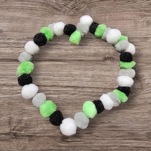 May include: A heart-shaped garland made of fluffy pom-poms in a variety of colors, including white, black, gray, and bright green. The pom-poms are arranged in a circular pattern, creating a decorative accent.