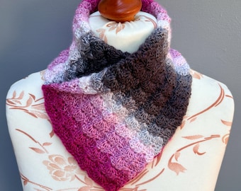 Puede incluir: Una bufanda de cuello tejida a ganchillo con un patrón de colores degradados que transita de magenta intenso y rosa a lavanda claro, gris y gris oscuro. El accesorio presenta un diseño de punto de concha texturizado en forma triangular.