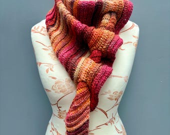 Puede incluir: Un pañuelo triangular tejido a ganchillo a mano con un degradado vibrante de colores cálidos que incluyen magenta intenso, naranja quemado, amarillo dorado y crema. El accesorio muestra un patrón de punto acanalado sobre un maniquí.