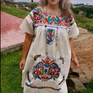 May include: Cream-colored embroidered dress with short sleeves, featuring vibrant floral patterns in red, orange, blue, and green. The dress has a round neckline and a long beaded necklace. The person is wearing black pants and red shoes.