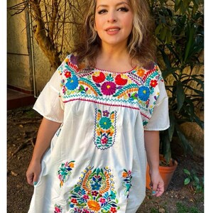 May include: White embroidered dress with colorful floral designs. The dress features short sleeves and a square neckline, adorned with vibrant red, blue, yellow, and purple flowers. The embroidery extends down the front of the dress, creating a festive and eye-catching look.