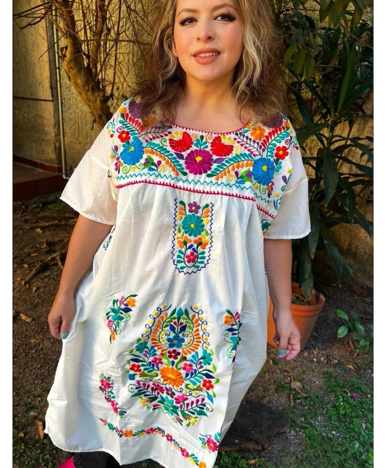 May include: White embroidered dress with colorful floral designs. The dress features short sleeves and a square neckline, adorned with vibrant red, blue, yellow, and purple flowers. The embroidery extends down the front of the dress, creating a festive and eye-catching look.
