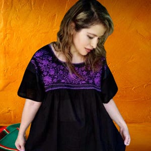 Puede incluir: Túnica negra bordada con flores moradas. La parte superior tiene mangas cortas y un escote redondo. El fondo es una pared naranja texturizada.