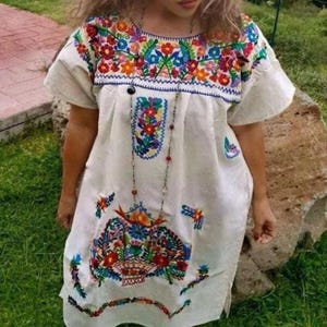 May include: A white, short-sleeved dress with vibrant floral embroidery in red, orange, blue, and green. The embroidery is concentrated around the neckline, chest, and hem. A long beaded necklace hangs down the front.