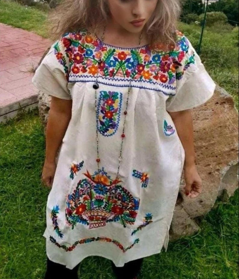 May include: A white, short-sleeved dress with vibrant floral embroidery in red, orange, blue, and green. The embroidery is concentrated around the neckline, chest, and hem. A long beaded necklace hangs down the front.