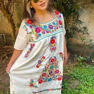 May include: A white embroidered dress with a scoop neckline and short sleeves. The dress features colorful floral embroidery in shades of red, blue, purple, and green. The dress is knee-length.