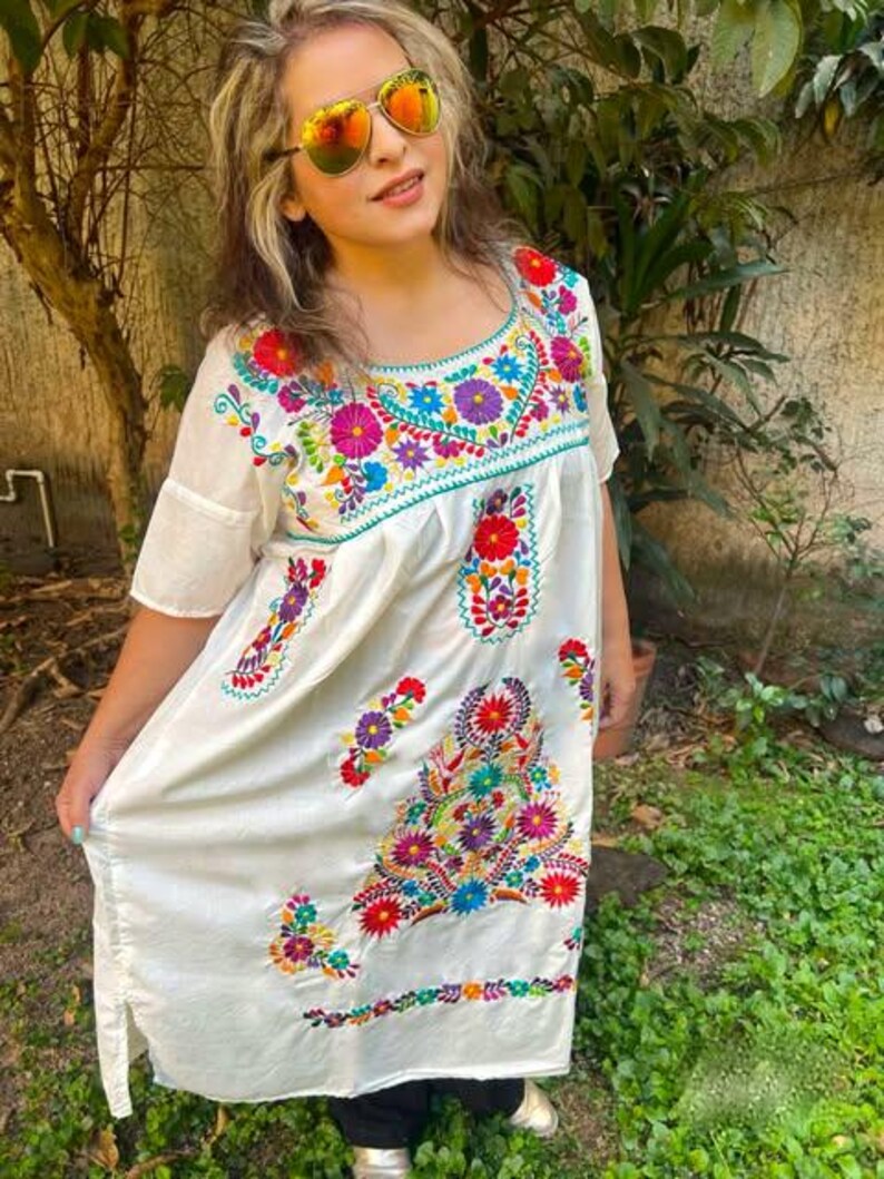 May include: A white embroidered dress with a scoop neckline and short sleeves. The dress features colorful floral embroidery in shades of red, blue, purple, and green. The dress is knee-length.