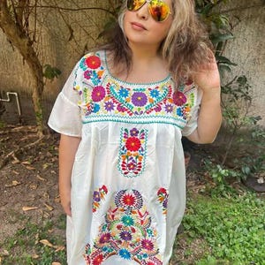 May include: White embroidered dress with colorful floral patterns. The dress features a round neckline and short sleeves. The embroidery includes red, pink, blue, and yellow flowers. The dress is knee-length.
