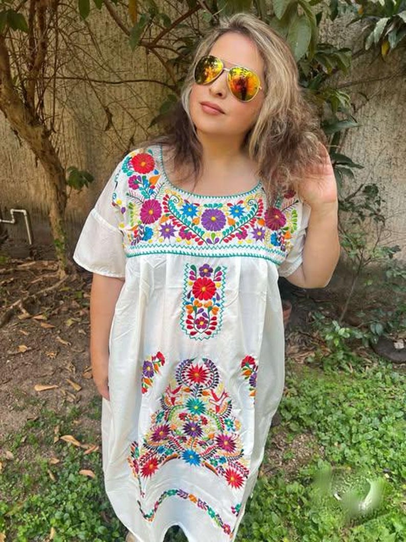 May include: White embroidered dress with colorful floral patterns. The dress features a round neckline and short sleeves. The embroidery includes red, pink, blue, and yellow flowers. The dress is knee-length.