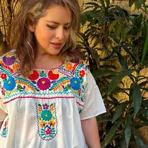 May include: White embroidered blouse with a vibrant floral design in shades of blue, red, yellow, orange, and purple. The blouse features short sleeves and a square neckline, showcasing a traditional Mexican style.