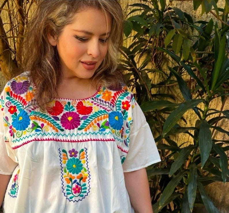May include: White embroidered blouse with a vibrant floral design in shades of blue, red, yellow, orange, and purple. The blouse features short sleeves and a square neckline, showcasing a traditional Mexican style.