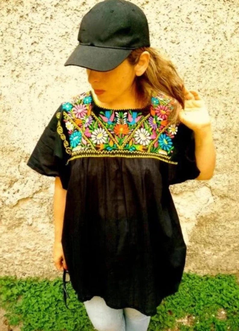 K&ouml;nnte beinhalten: Eine schwarze, bestickte mexikanische Bluse mit einem floralen Design in leuchtenden Farben. Die Bluse hat kurze &Auml;rmel und einen runden Ausschnitt. Auf dem Kopf wird eine schwarze Baseballkappe getragen.