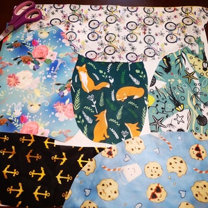 Puede incluir: Muestras de tela variadas con diversos patrones, incluyendo bicicletas, zorros, anclas y galletas. Unas tijeras moradas están colocadas en la esquina superior izquierda. Las telas están cortadas en formas irregulares, sugiriendo un proyecto de manualidades.