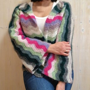 May include: A colourful crocheted cardigan with a wavy striped pattern in shades of pink, green, purple, and white. The cardigan has long sleeves and a relaxed fit.