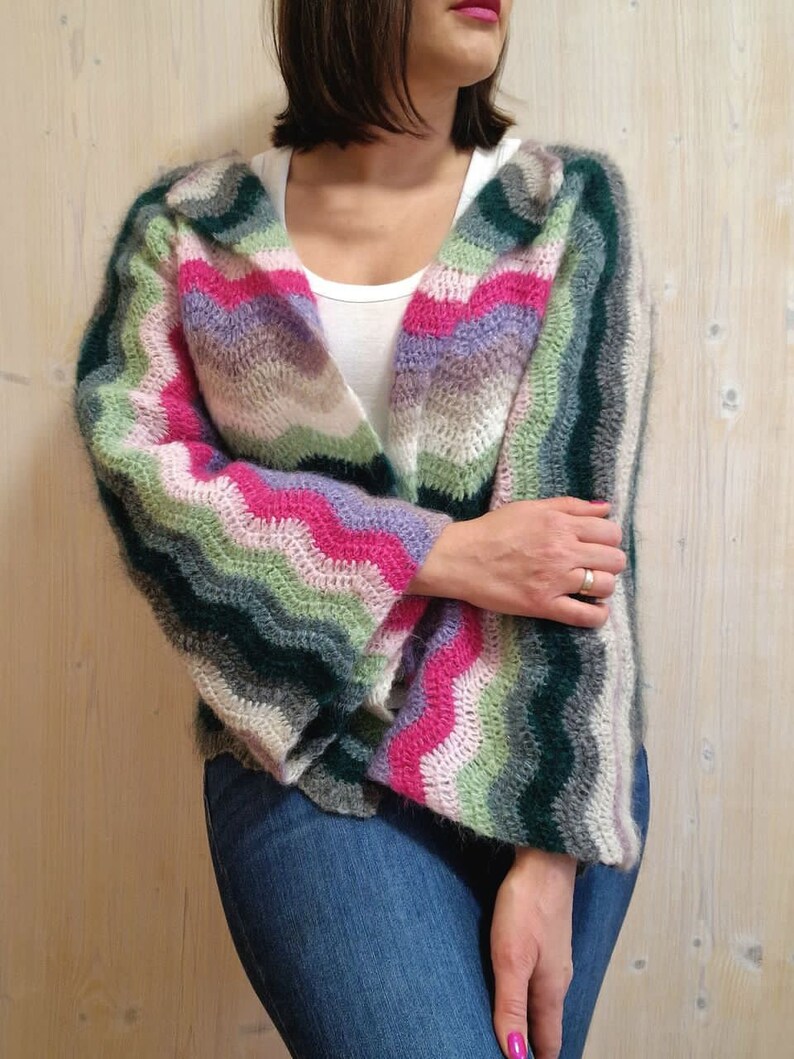 May include: A colourful crocheted cardigan with a wavy striped pattern in shades of pink, green, purple, and white. The cardigan has long sleeves and a relaxed fit.