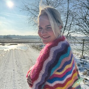 May include: A woman wearing a colourful, striped, crocheted sweater with a wavy pattern. The sweater is made of a soft, fuzzy yarn and features a variety of colours, including pink, purple, blue, green, yellow, and orange. The woman is standing in a snowy field with a bright blue sky in the background.