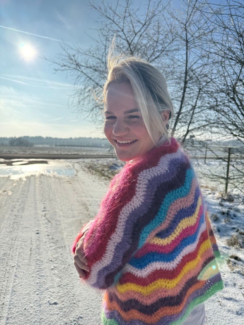 May include: A woman wearing a colourful, striped, crocheted sweater with a wavy pattern. The sweater is made of a soft, fuzzy yarn and features a variety of colours, including pink, purple, blue, green, yellow, and orange. The woman is standing in a snowy field with a bright blue sky in the background.