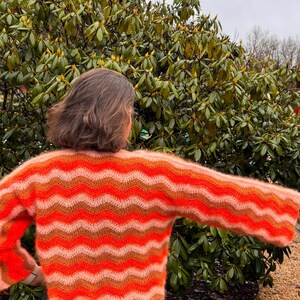 May include: A woman wearing a bright orange, pink and brown striped crocheted sweater with long sleeves. The sweater has a wavy pattern.