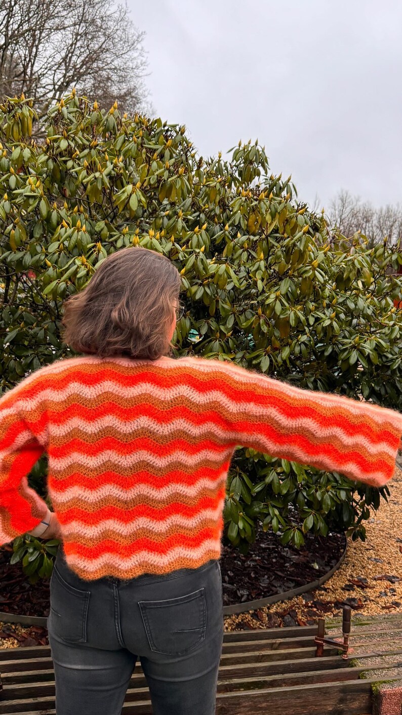 May include: A woman wearing a bright orange, pink and brown striped crocheted sweater with long sleeves. The sweater has a wavy pattern.
