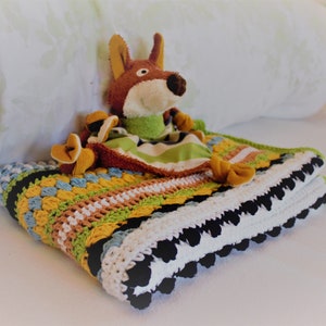 Könnte beinhalten: Eine bunte gehäkelte Babydecke mit einem schwarzen und weißen Rand und einem braunen und grünen Fuchs-Plüschtier.