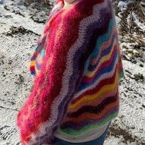 May include: A colourful, fuzzy, crocheted sweater with a wavy striped pattern in shades of pink, purple, blue, green, yellow, and orange. The sweater is worn over a white shirt and blue jeans.
