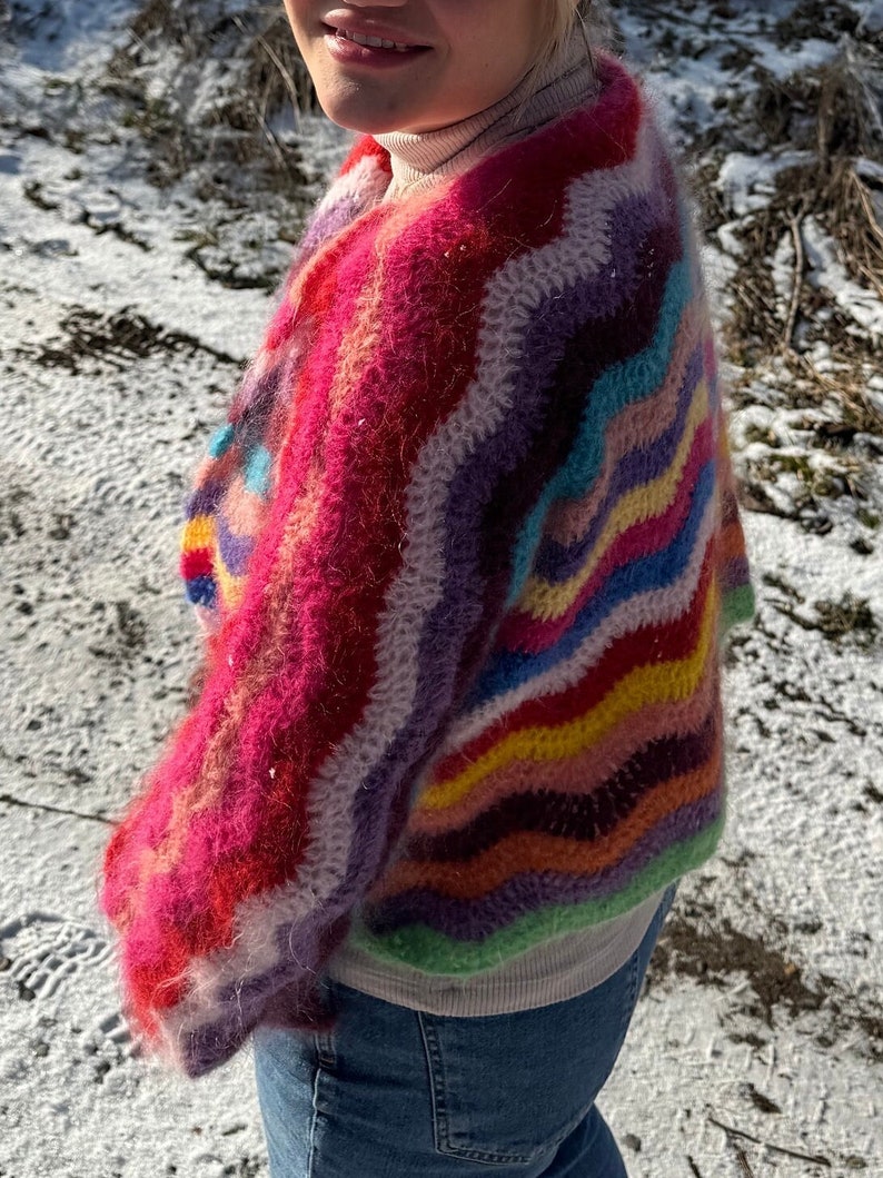May include: A colourful, fuzzy, crocheted sweater with a wavy striped pattern in shades of pink, purple, blue, green, yellow, and orange. The sweater is worn over a white shirt and blue jeans.
